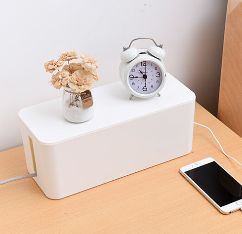 A white rectangular cable management box on a light wood table, styled with a small vase of dried flowers and an alarm clock on top, with a smartphone charging from a cable plugged into the side of the box.