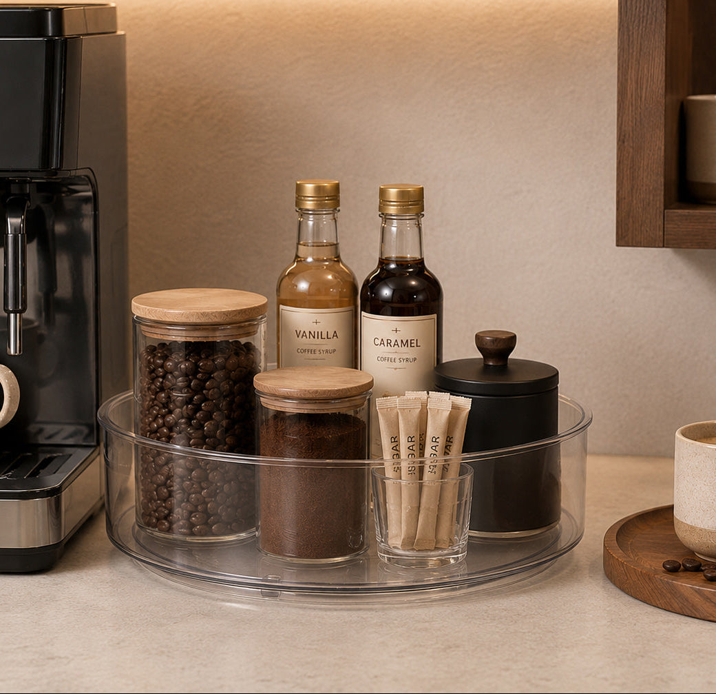 A countertop coffee station setup with a black espresso machine on the left, a clear round tray holding glass jars of coffee beans and syrup, two small bottles labeled Vanilla and Caramel, and a black lidded container. A beige mug sits on the counter, with a wooden shelf and warm ambient lighting in the background.