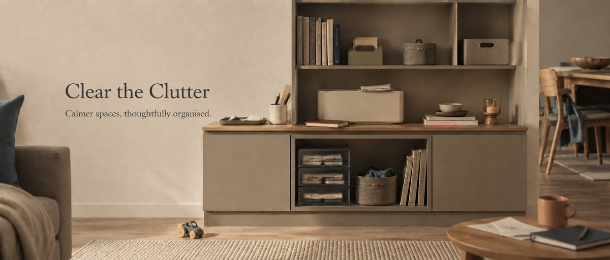 A styled interior scene showing a tall storage cabinet and shelving unit in a warm neutral room. The cabinet has a taupe-gray body with a wood top and open shelves holding books, boxes, and small decor items.