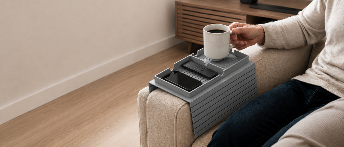 A gray plastic armrest organizer tray resting on a beige armchair, holding a white mug, with a person seated beside it in a living room setting.