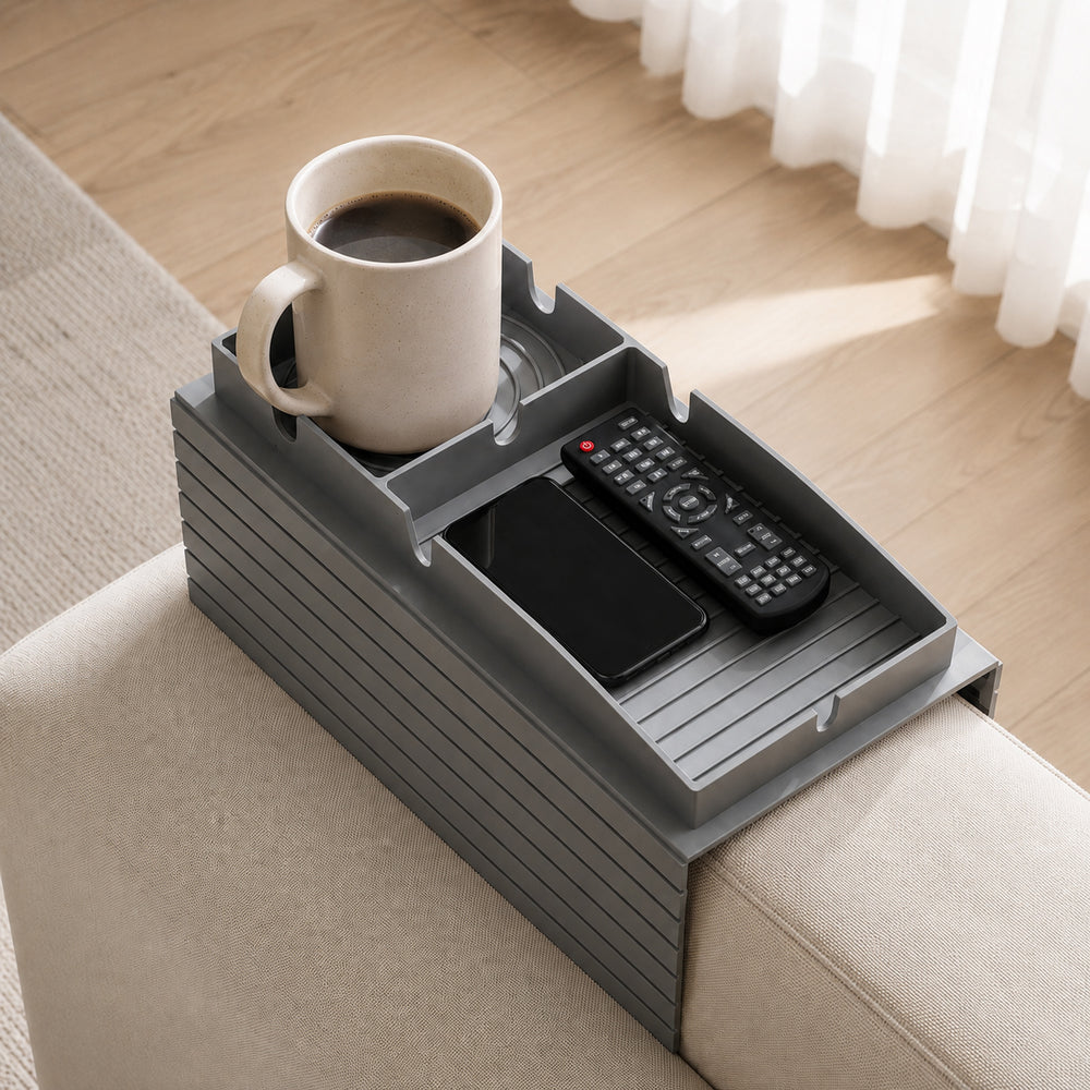 A gray plastic armrest organizer tray resting on a beige armchair in a bright living room. The tray holds a white mug filled with black coffee, a black remote control, and a black smartphone.